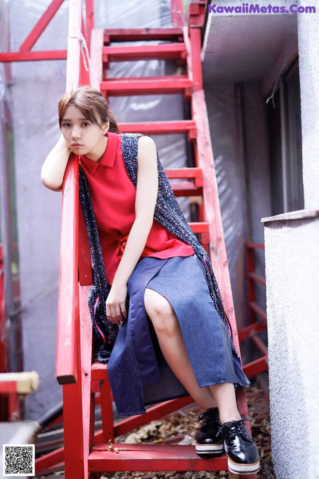 A woman sitting on a red ladder in a red shirt and blue skirt.